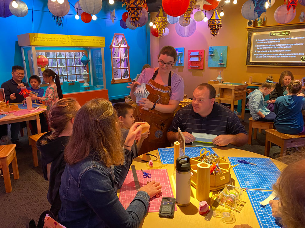 Children and grown-ups sitting around a table. A museum staff member is holding up a paper lantern for them to see.